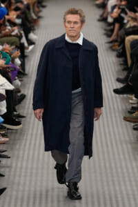 Male model walking a fashion runway in a navy overcoat, dark sweater, light gray pants, and black boots; seated audience on both sides.