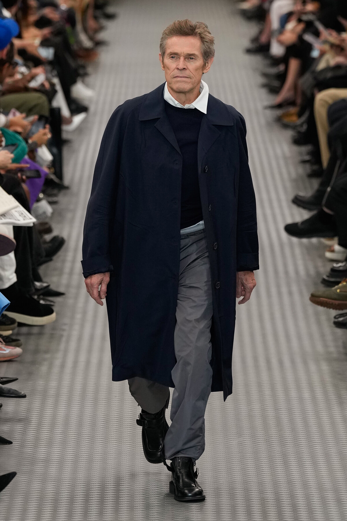 Male model walking a fashion runway in a navy overcoat, dark sweater, light gray pants, and black boots; seated audience on both sides.
