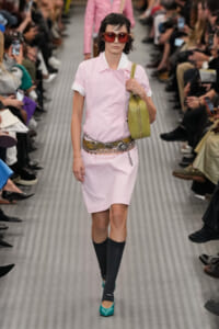 Model walking a runway in a pastel pink dress with rolled sleeves, oversized red sunglasses, a wide decorative belt, and teal heels, carrying a lime-green handbag.