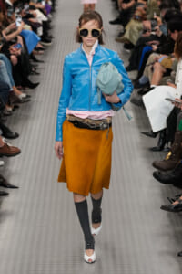 Model walks the runway in a shiny blue cropped jacket, pink top, mustard skirt, and white heels with oversized sunglasses, holding a teal bag.