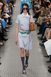 Runway model in a light-blue shirt dress with a teal belt, brown tote, and oversized purple sunglasses walking down the catwalk.