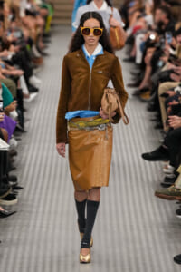 Model on a runway wearing a brown suede zip-front jacket over a blue shirt, a metallic gold skirt, oversized yellow sunglasses, knee-high socks, gold shoes, and a beige handbag