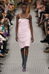 Model walks a fashion runway in a pastel pink dress with white straps, gray knee-high socks, and purple heels; audience lines the catwalk in the background.