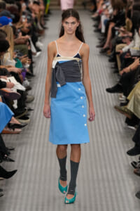 Model walking a fashion runway in a light blue knee-length skirt with white buttons, a dark gray wrap, and teal open-toe heels with gray socks.