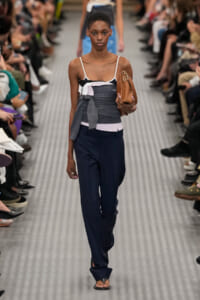 Black female model walking a fashion runway in a gray layered top with white straps, navy trousers, black sandals, and a tan handbag tucked under her arm.