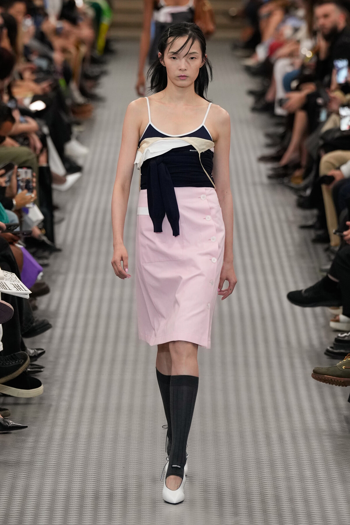 Model walks the runway in a pale pink knee-length skirt with white buttons, paired with a navy and cream top and white heels.