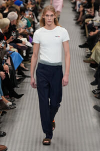 Male model walks a fashion-show runway in a white T-shirt and navy trousers, brown sandals, with a seated audience on both sides.