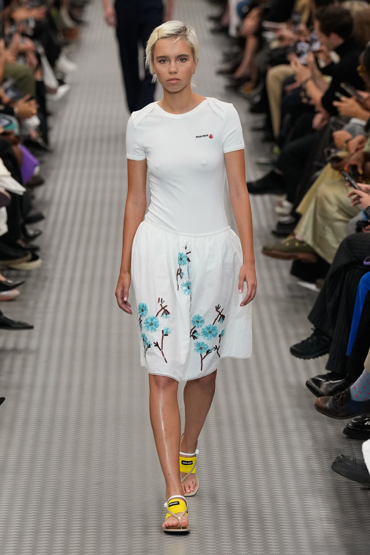 Model walking the runway in a white top and embroidered white skirt with blue flowers; audience sits along the catwalk behind her.