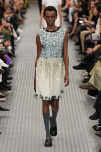 Model on a runway wearing a sleeveless silver sequin top and a pale gradient skirt with circular appliqués, paired with gray knee-high socks and black loafers.
