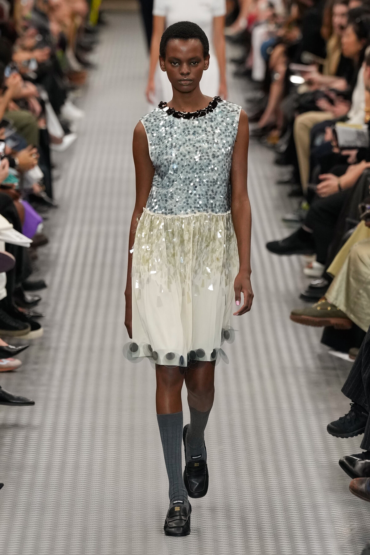 Model on a runway wearing a sleeveless silver sequin top and a pale gradient skirt with circular appliqués, paired with gray knee-high socks and black loafers.