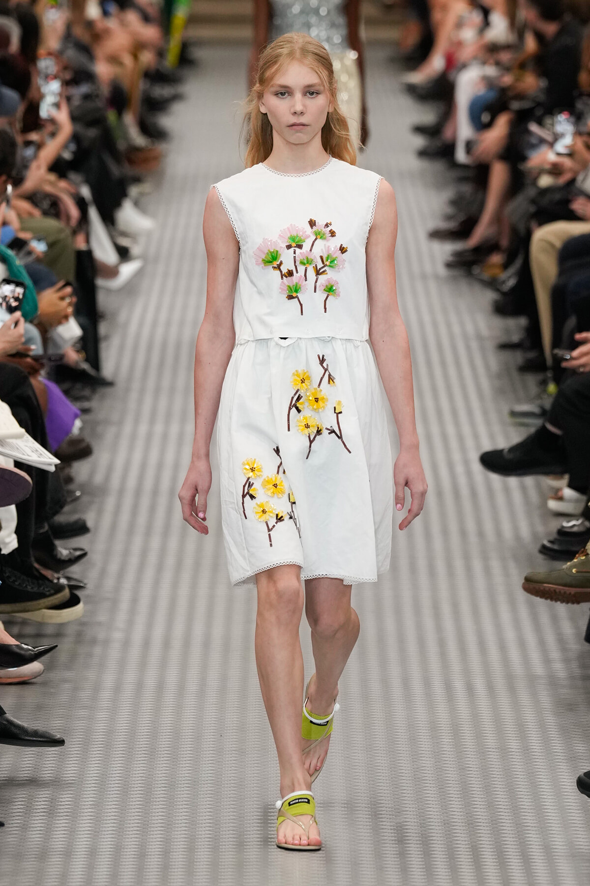 Model walking a runway in a white sleeveless dress with embroidered pink and green flowers, yellow accents, and brown branches.