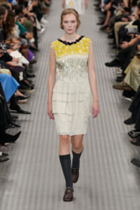 Runway model walks in a white tiered dress with a yellow sequin bodice and black beaded neckline, paired with dark knee-high socks and brown loafers.