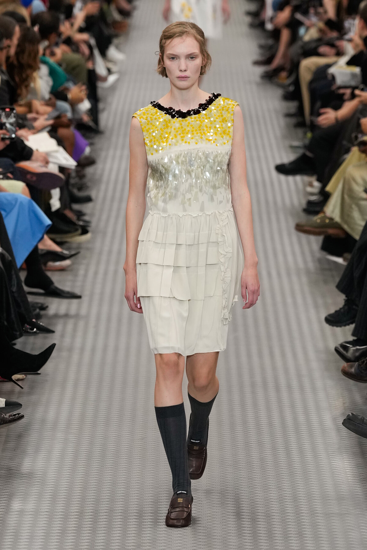 Runway model walks in a white tiered dress with a yellow sequin bodice and black beaded neckline, paired with dark knee-high socks and brown loafers.
