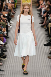 Model walks a fashion runway in a white fitted top and pleated white midi skirt, with brown shoulder straps and yellow-black sandals.