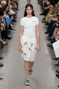 Model walks the runway in a white short-sleeve top and knee-length skirt with pink floral embroidery, turquoise sandals, audience on both sides.