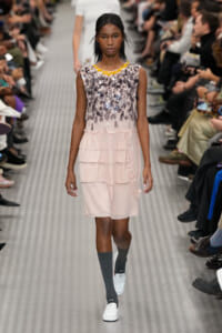 Model walks the runway in a light pink, sleeveless dress with a layered, ruffled skirt and a sequined gray-to-black bodice accented by a yellow neckline, paired with gray knee-high socks and white sneakers.