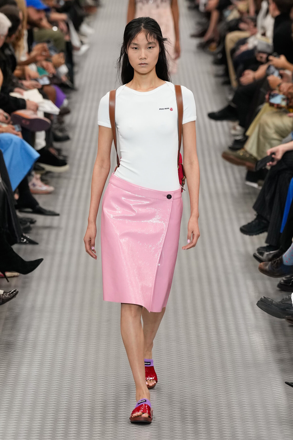 Model walks a fashion runway wearing a white fitted tee and glossy pink knee-length skirt with brown shoulder straps for a Miu Miu show, audience seated on both sides.