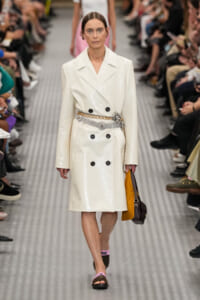 Female model on a runway wearing a white shiny double-breasted trench coat with black buttons, a metallic belt, and carrying a brown handbag.