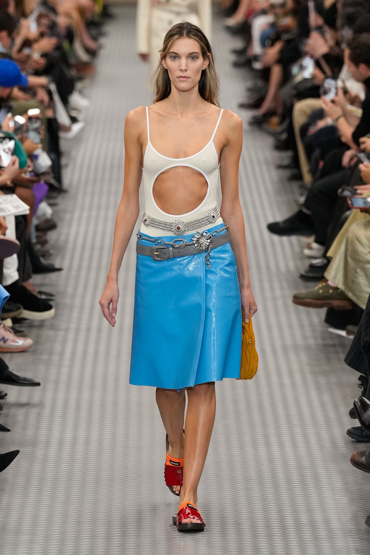 Model walking a runway in a white cutout tank top, bright blue skirt, and orange slide sandals, audience on both sides