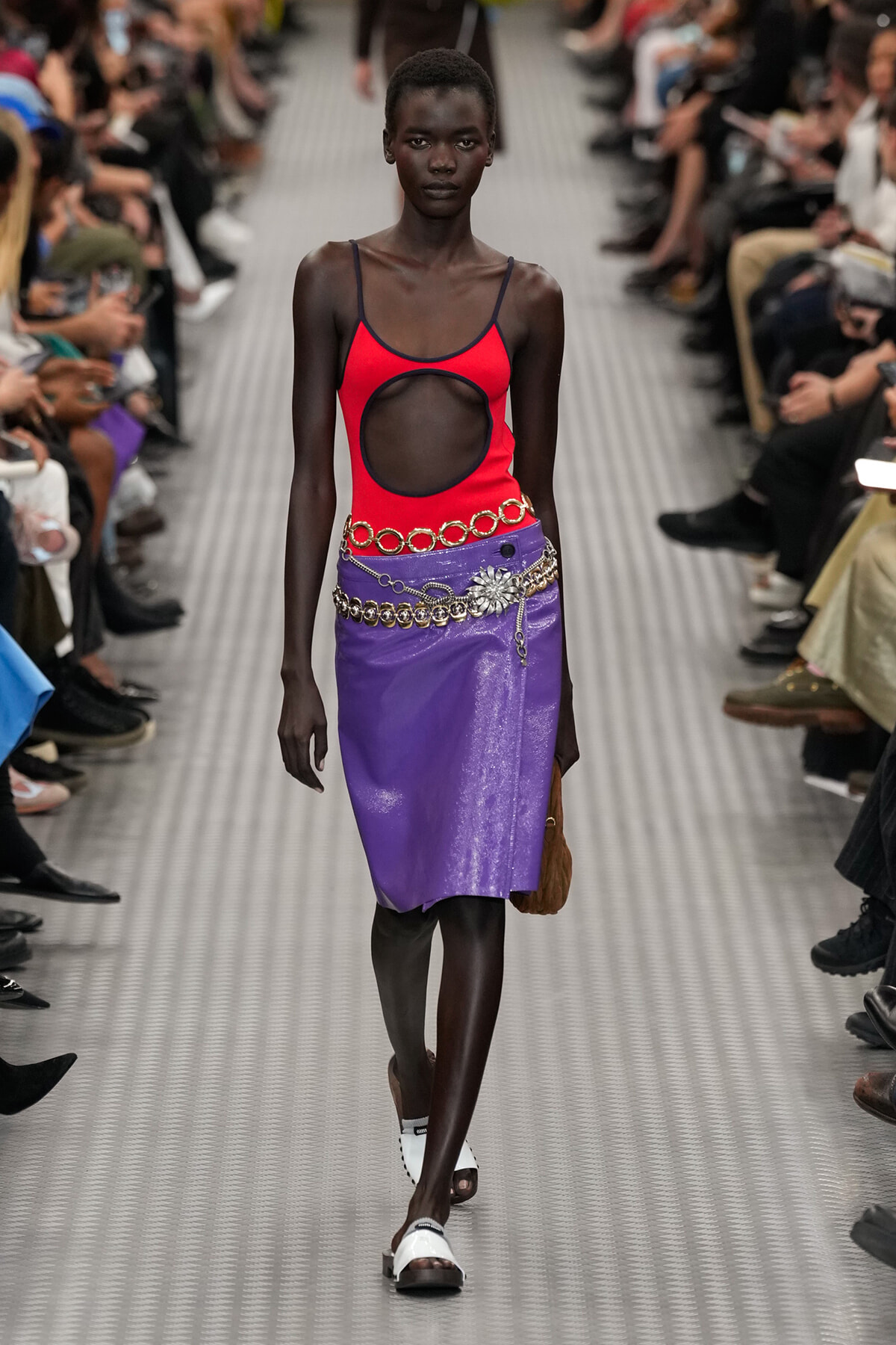 Model walking the runway in a red cut-out tank top and shiny purple skirt with a silver chain belt, audience on both sides of the catwalk.