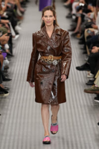 Model walking the runway in a glossy brown trench coat with a tan belt and decorative chain belt, paired with pink slides on a metallic catwalk