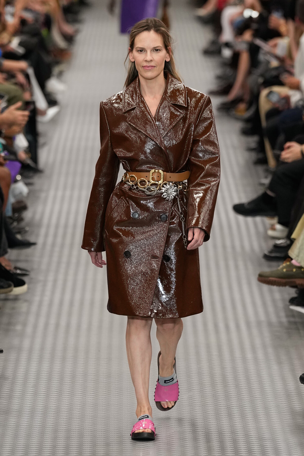 Model walking the runway in a glossy brown trench coat with a tan belt and decorative chain belt, paired with pink slides on a metallic catwalk