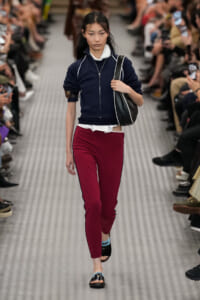 Model walks a fashion show wearing a navy zip-up cropped jacket, white shirt, burgundy pants with black piping, and black sandals, carrying a black shoulder bag.