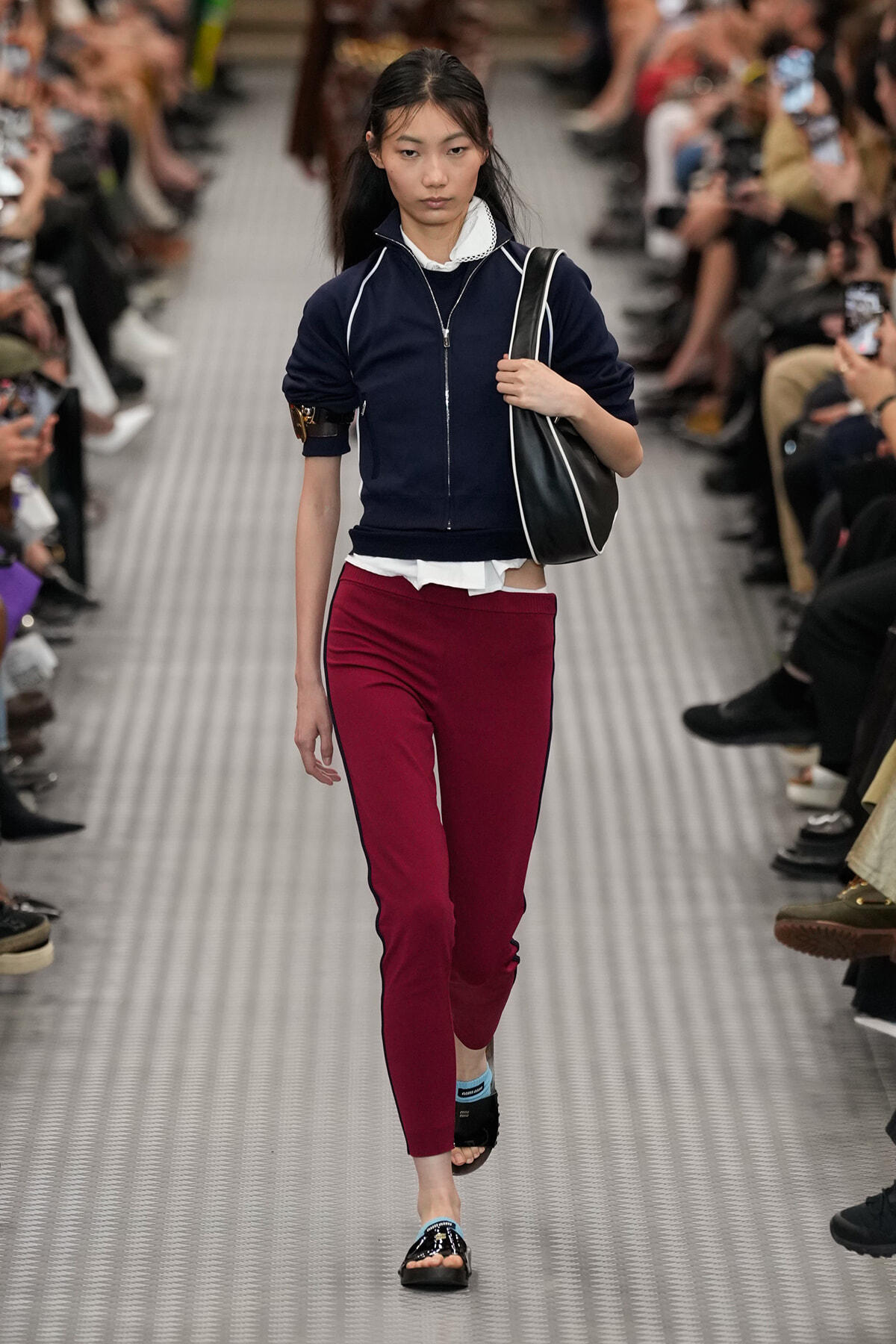 Model walks a fashion show wearing a navy zip-up cropped jacket, white shirt, burgundy pants with black piping, and black sandals, carrying a black shoulder bag.