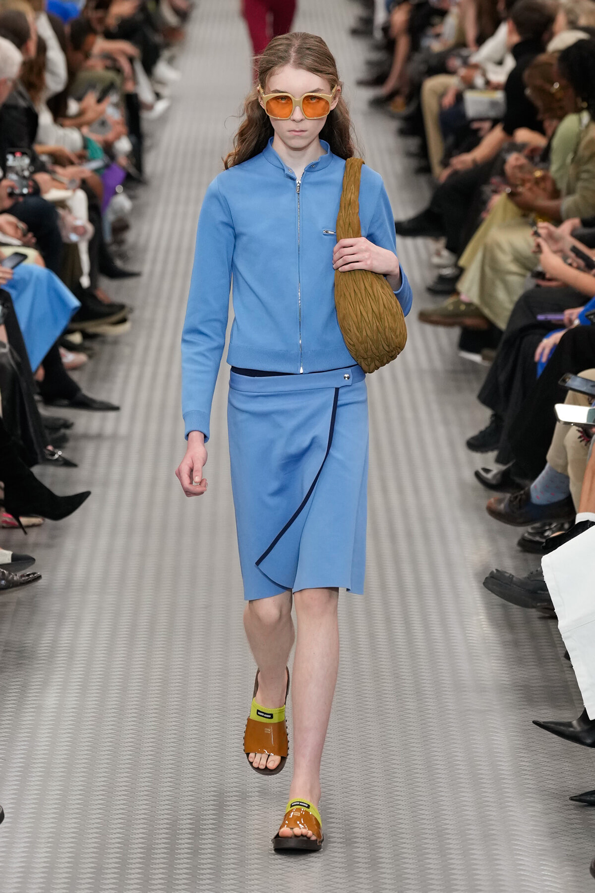 Model walks the runway in a light blue zip-front jacket and matching knee-length skirt, wearing orange sunglasses and carrying a brown woven bag over the shoulder, with lime-green-accented sandals.
