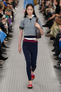 Model walks the runway wearing a light gray zip jacket, navy leggings with white side stripes, burgundy waistband, red slide sandals, and a brown shoulder bag.