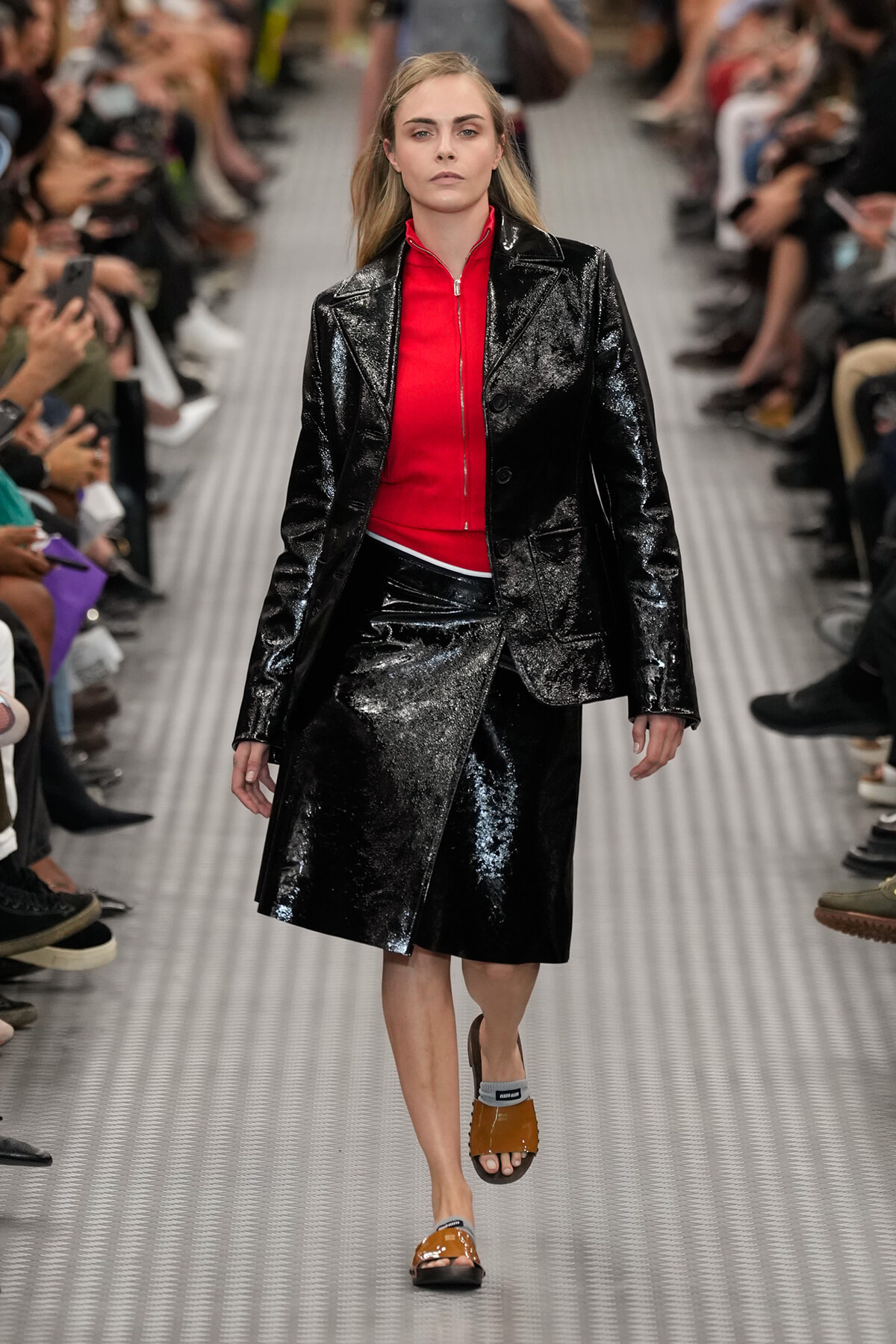 Model walking the runway in a shiny black patent blazer and matching skirt with a bright red zip-top underneath, audience on sides.