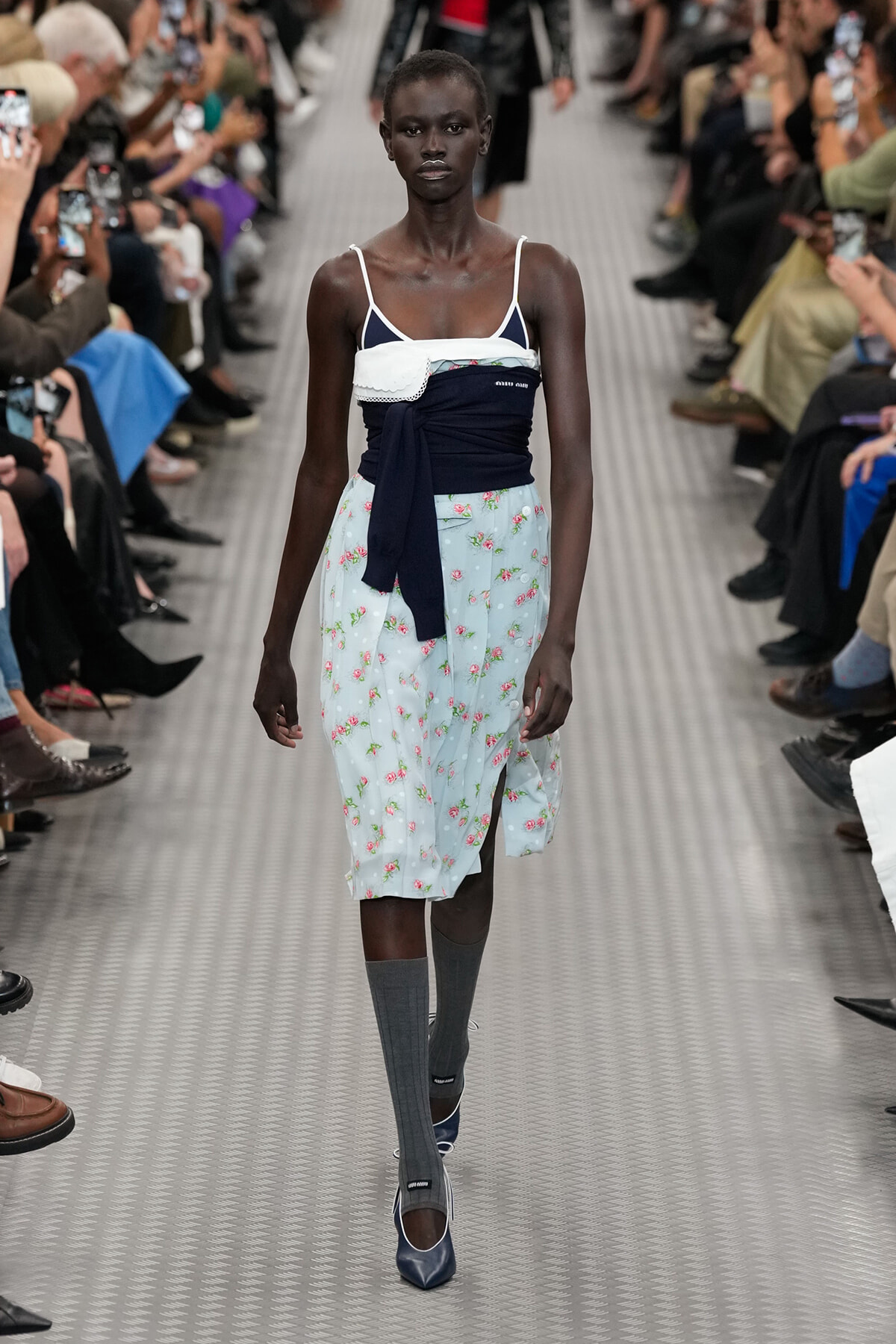 Model walking a runway in a light blue floral midi skirt with a navy sash, white top, gray knee-high socks, and blue pumps.