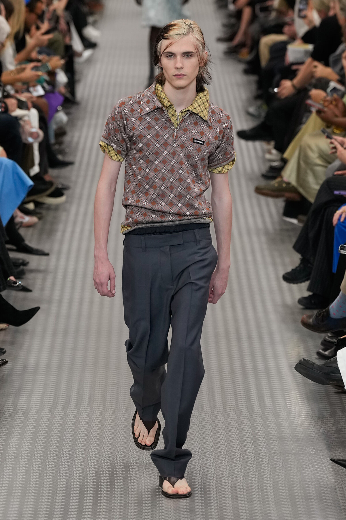 Model walking a fashion runway in a brown plaid polo with yellow collar and rolled sleeves, paired with gray trousers and flip-flops.