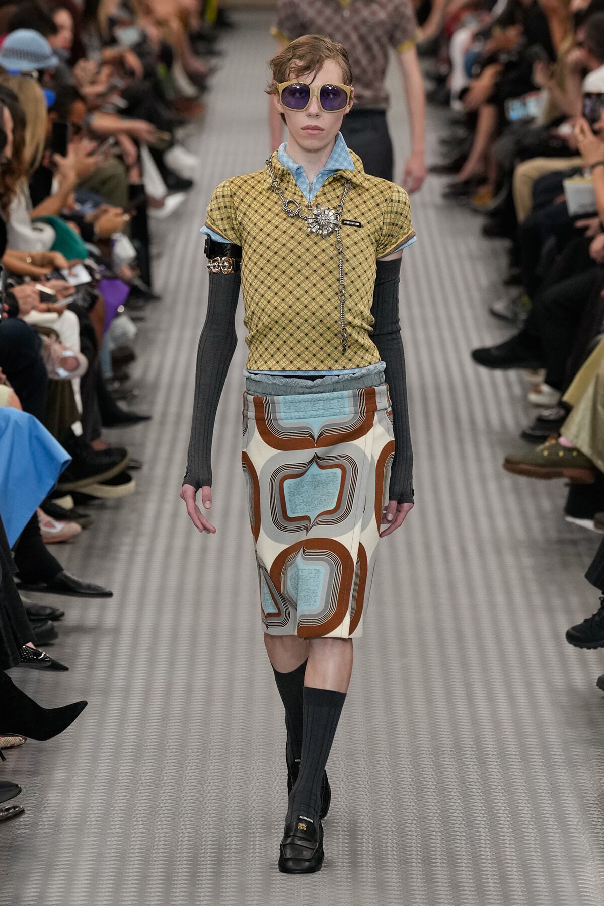 Model walks the runway in a mustard-yellow, checkered short-sleeve top, layered with denim collar, long black arm-warmers, and a retro abstract-print pencil skirt; oversized sunglasses and accessories complete the look.