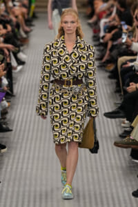 Model walks runway in a black‑white geometric swirl patterned coat dress with lime accents, cinched with a wide brown belt and a silver floral chain accessory, carrying a brown/black bag and wearing mint sneakers with yellow laces.
