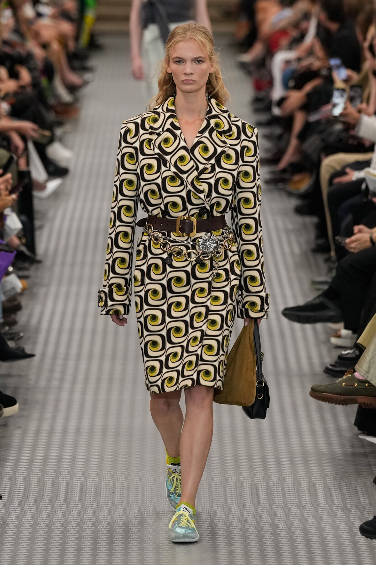 Model walks runway in a black‑white geometric swirl patterned coat dress with lime accents, cinched with a wide brown belt and a silver floral chain accessory, carrying a brown/black bag and wearing mint sneakers with yellow laces.