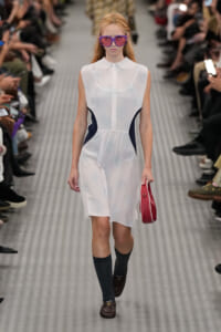 Runway model wearing a white sleeveless dress with navy side panels, pink sunglasses, and a red clutch.