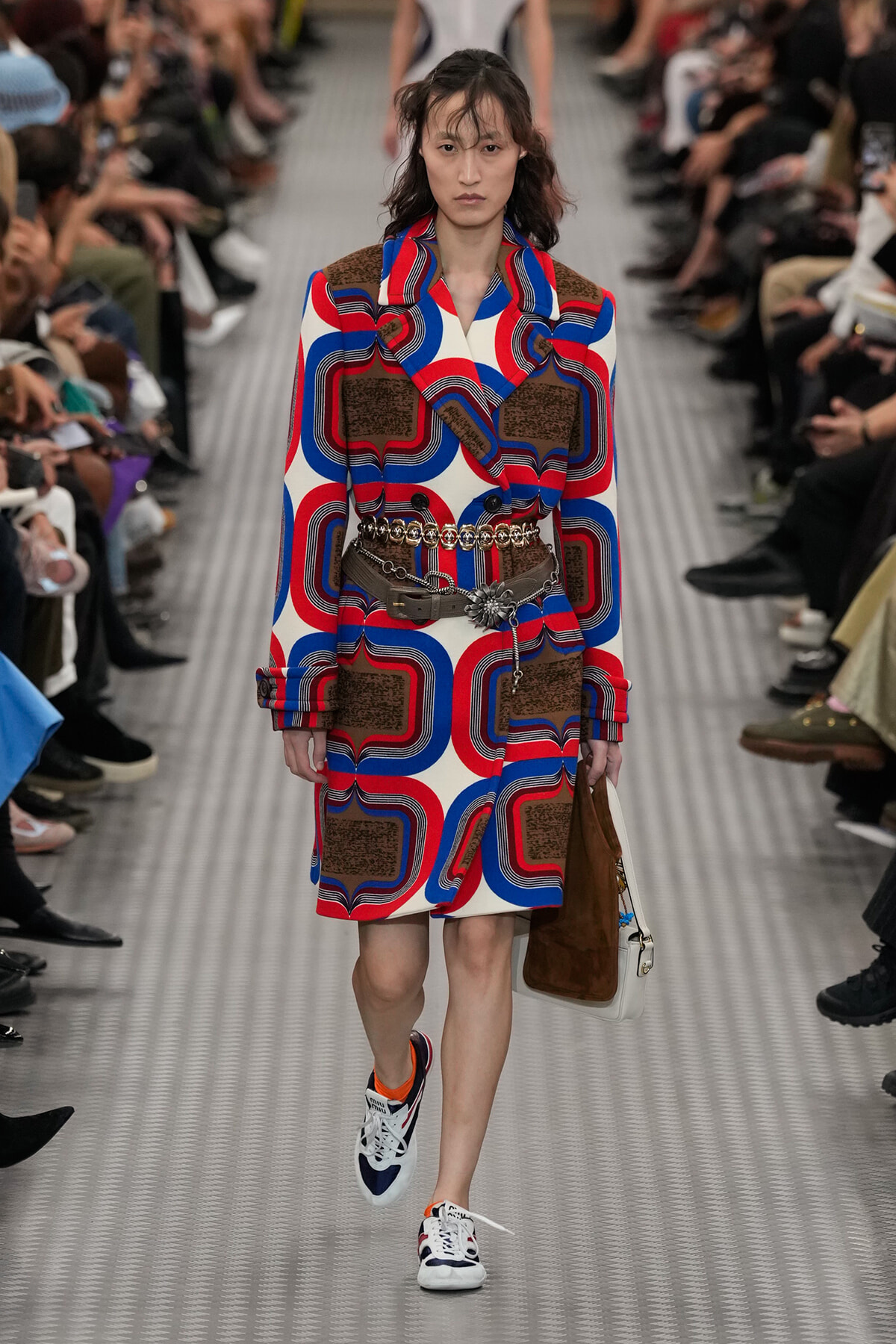 Model walks a runway in a bold red, blue, and brown geometric coat with a wide belt and a brown suede bag.
