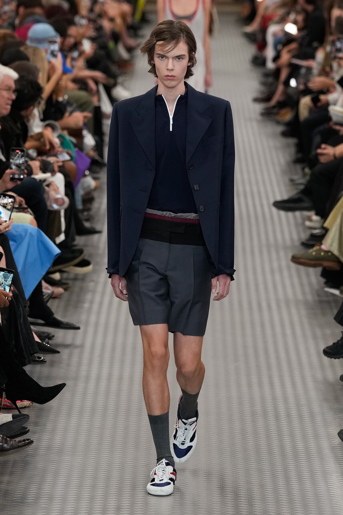 Model walks a fashion runway in a navy blazer, navy zip-neck top, gray shorts, and color-block sneakers with dark socks; audience on both sides.