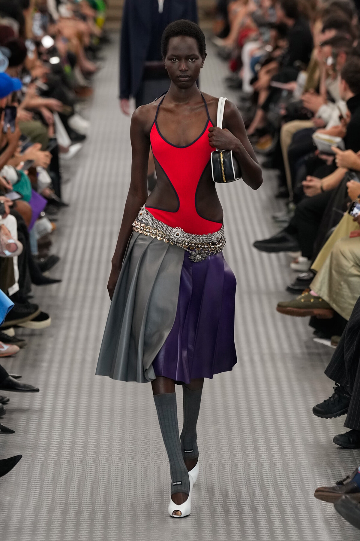 Runway model in a red cut-out swimsuit top with a silver belt and color-block gray-to-purple skirt, gray socks, and white heels.