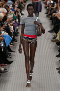 Runway model walking a fashion show in a light blue top, red waistband, chain belt, dark bikini bottoms, white sandals, and a brown shoulder bag.