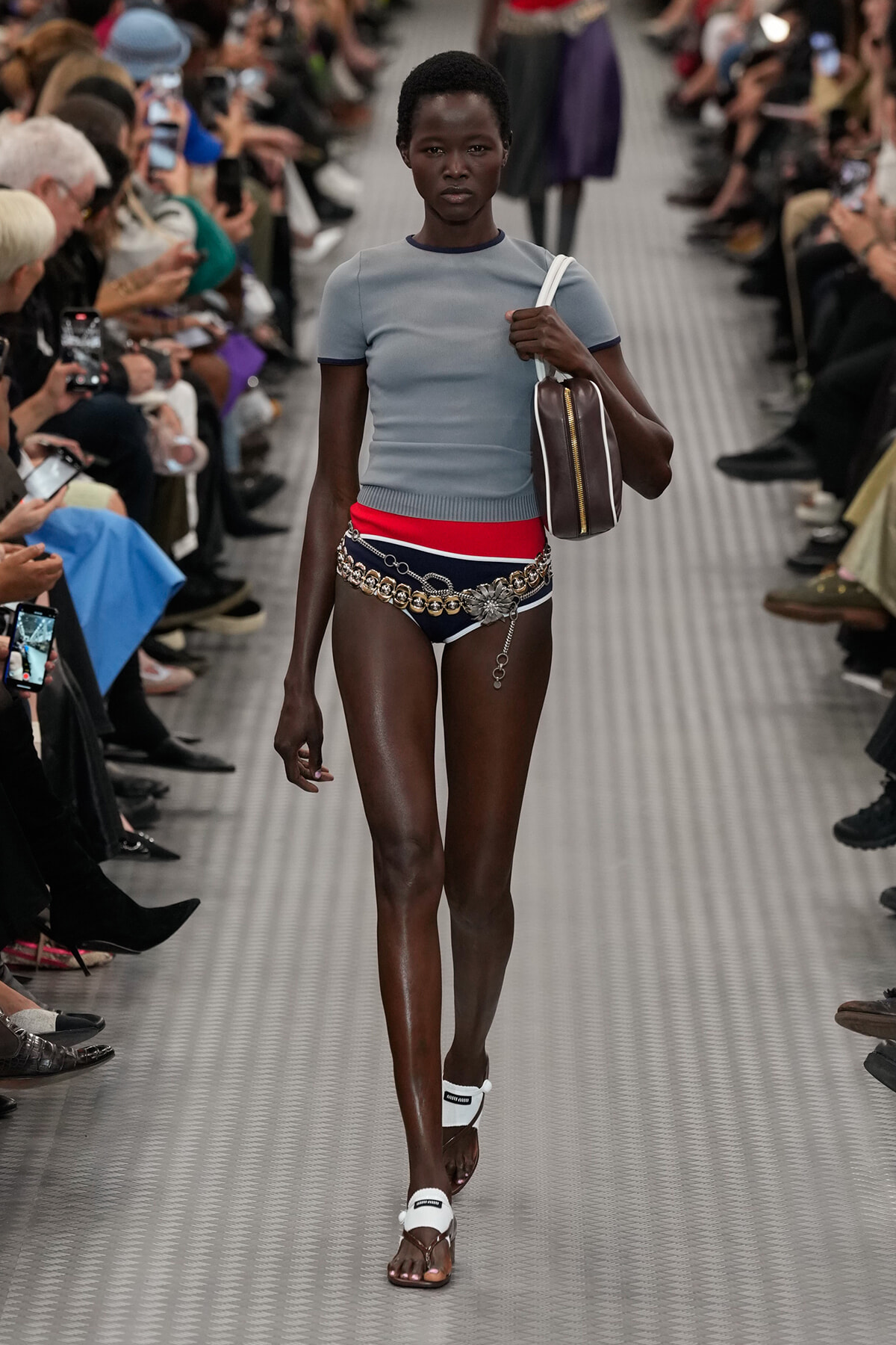 Runway model walking a fashion show in a light blue top, red waistband, chain belt, dark bikini bottoms, white sandals, and a brown shoulder bag.