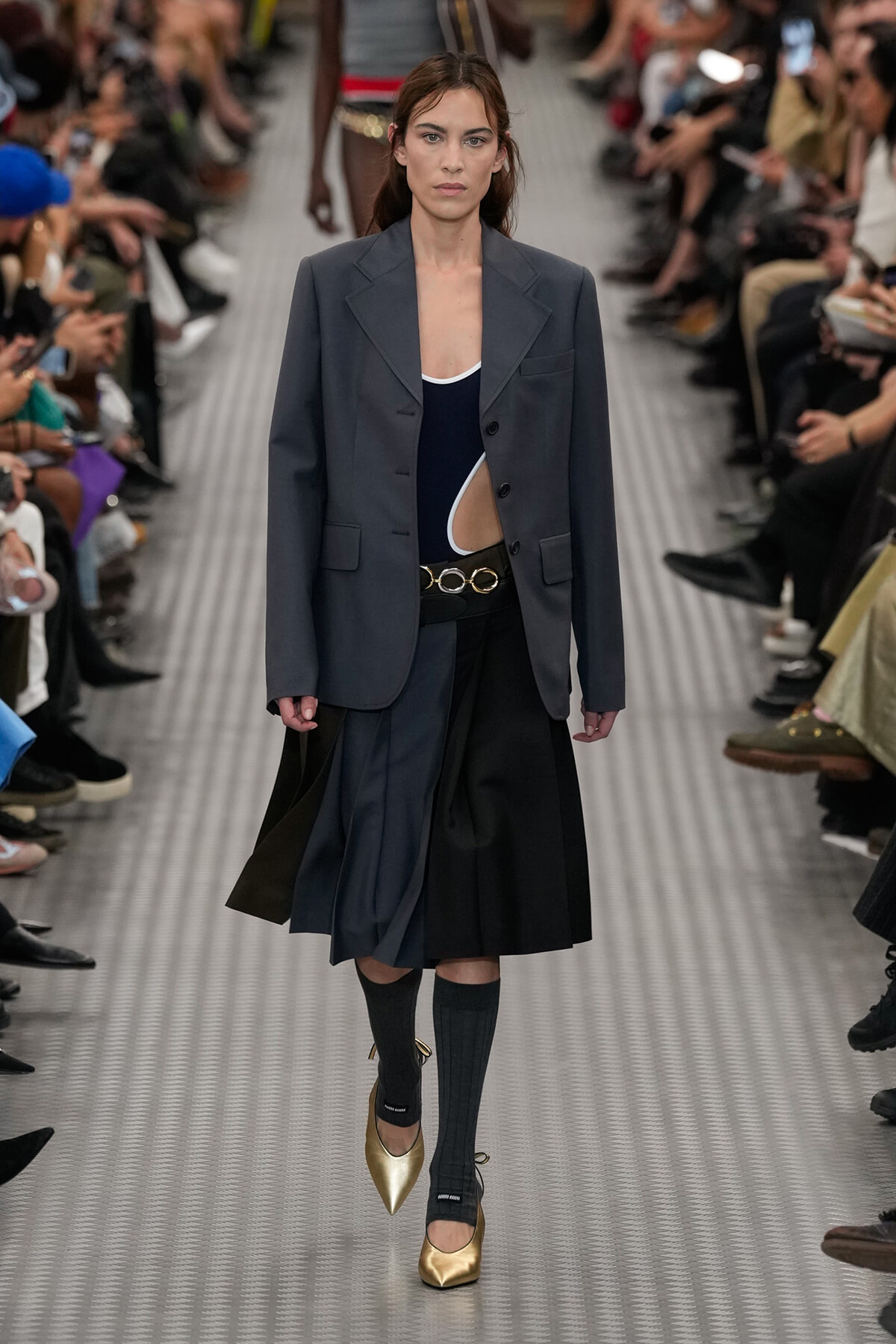 Model walking a runway in an oversized dark gray blazer over a navy cutout top, paired with a color-blocked pleated skirt and gold pointed-toe heels, black socks, and a circular-buckle belt.