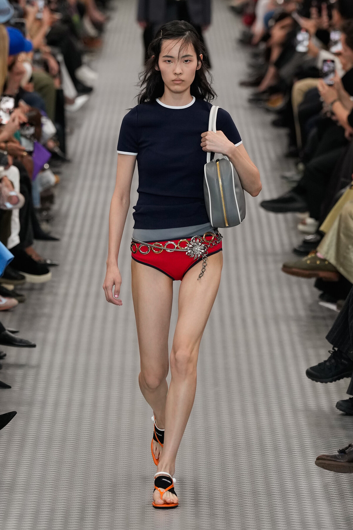 Model on a runway wearing a navy short-sleeve top, red bikini bottoms with a chain belt, orange sandals, and a gray bag over the shoulder.