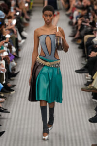 Model walking a runway in a strapless gray top with cutouts and a teal and black color-blocked skirt with a jeweled belt. A small handbag rests on her shoulder.