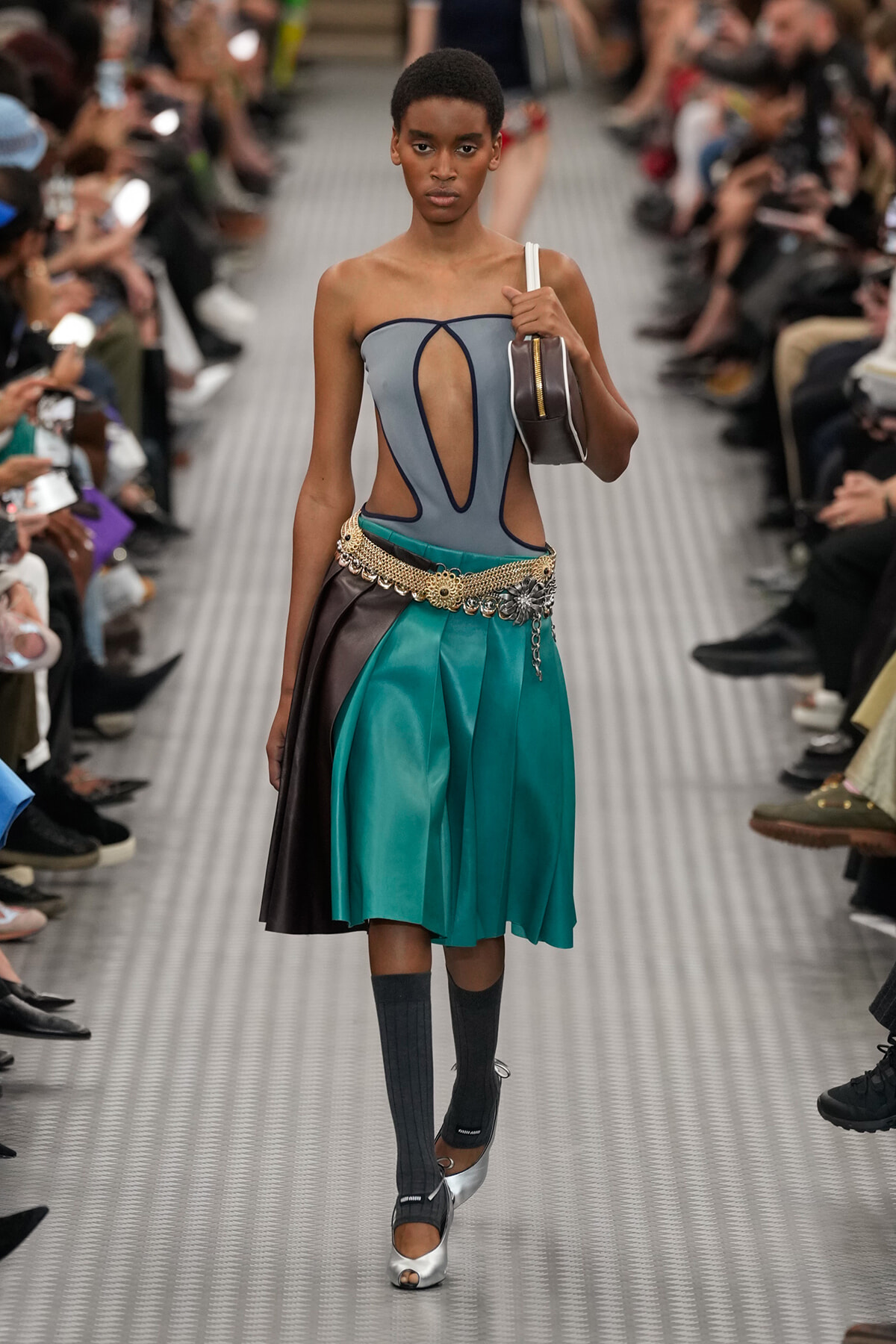 Model walking a runway in a strapless gray top with cutouts and a teal and black color-blocked skirt with a jeweled belt. A small handbag rests on her shoulder.