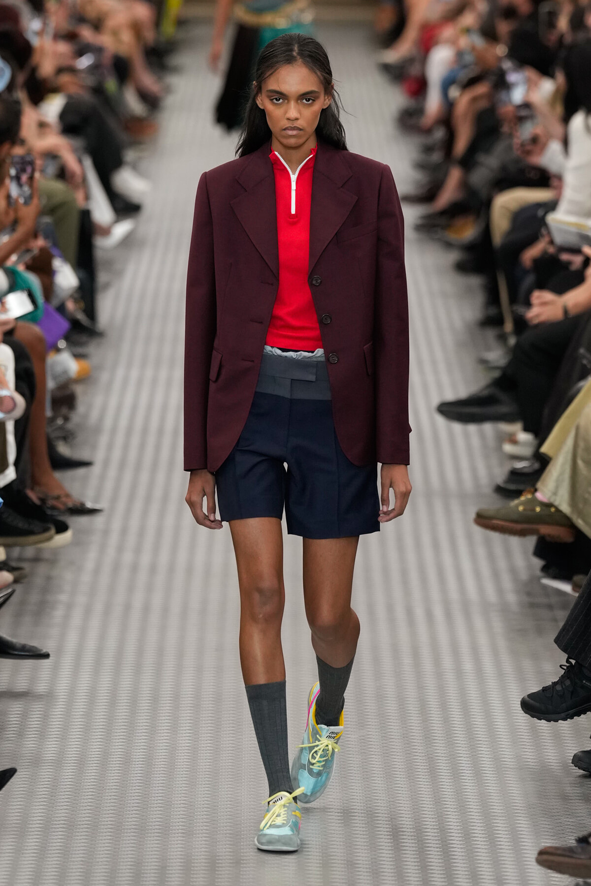 Model walking a runway in a burgundy blazer, red zip-neck top, navy shorts, gray knee-high socks, and pastel sneakers.