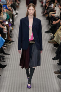 Model walking the runway in a navy blazer over a pink cutout bodysuit, with a color-blocked skirt and purple heels.