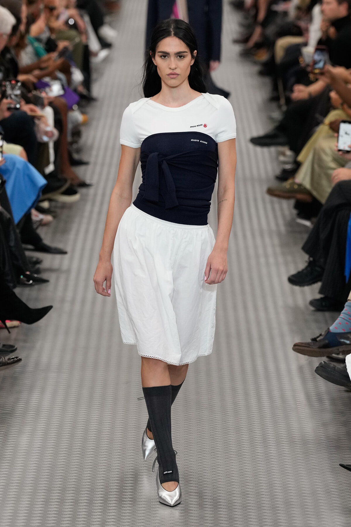 Model walking a fashion runway in a white short-sleeve top with a navy wrapped bandeau, white A-line skirt, black knee-high socks, and silver heels.