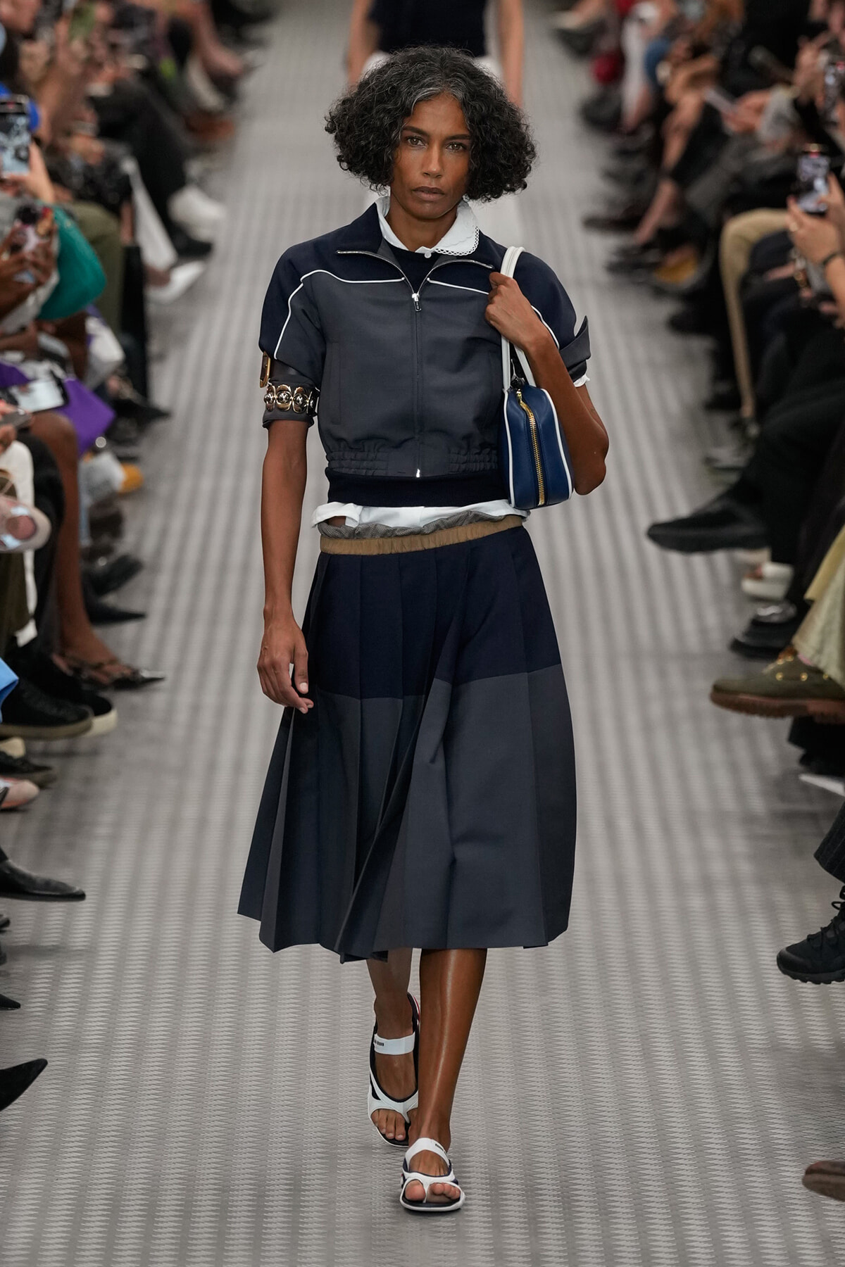 Female model walks a fashion runway in a navy cropped jacket with white piping, white shirt underneath, and a pleated midi skirt, carrying a blue handbag.